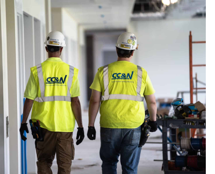 Two workers in hard hats and safety vests walking down a hallway at a construction site.