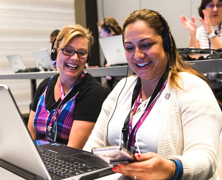 Two women at a conference smiling while looking at their laptops and wearing headsets.